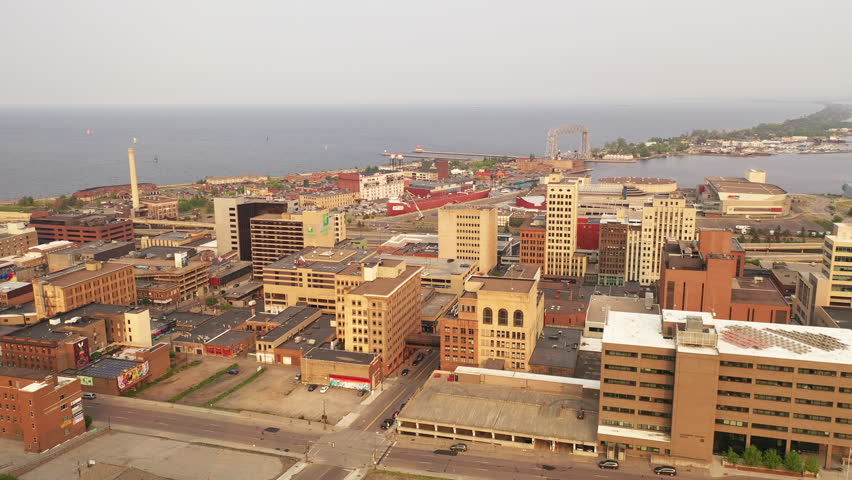 Aerial drone view of downtown Duluth Minnesota United States cityscape skyline at spring summer sunset. Beautiful birds eye footage captures the glowing urban skyline with high rise buildings harbor waterfront and dramatic orange sunset light over Lake Superior. Shot in crisp 4K this dynamic clip showcases the vibrant evening atmosphere of this iconic Great Lakes city. Perfect for travel documentaries Duluth tourism promotions cityscape footage sunset aerials urban lifestyle content or stock videos about Minnesota cities and Lake Superior scenery.