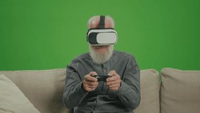 Green Screen. An Old Man With VR Glasses Plays Computer Games With a Joystick, Sitting on a Sofa. An Elderly Man Plays PlayStation and Wins. Tech Nostalgia and Generational Divide.
