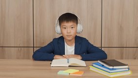 Distance learning, asian teen boy in a wireless headphones sitting on the desk in living room and study at home, homeschooling online by video call, the boy are listening attentively to the teacher.