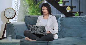 Beautiful Indian female entrepreneur sitting on comfortable sofa project task studying with laptop at indoor home. Professional business woman typing email browse on computer keyboard looking screen