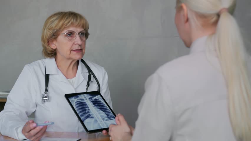 An elderly woman general practitioner or therapist or pulmonologist shows a female patient the results of a chest X-ray on an electronic tablet. High quality 4k footage Royalty-Free Stock Footage #1105450435