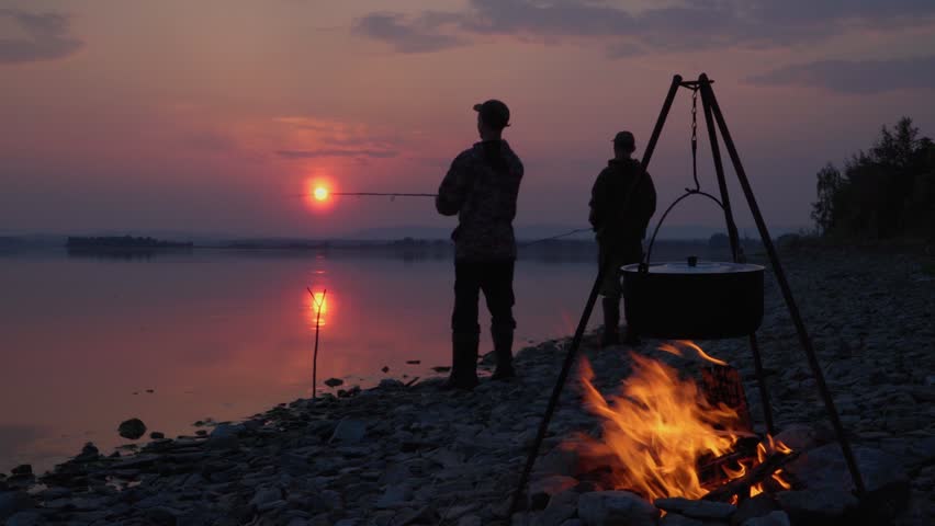 Friends at dusk at sunset are fishing on the shore of the lake. The fire is burning. Food is cooked on a campfire in a tourist cauldron.