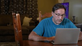 A man in his home dining room video chats on his laptop, smiling and waving to the screen. Complimentary reverse over the shoulder (OTS) shot available.  	