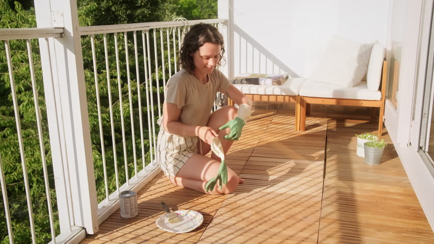Young  curly woman puts on rubber gloves  restoring floor decking on her terrace. Female  using a brush applies wood stain on the wooden floor on the balcony. DIY Royalty-Free Stock Footage #1105457867