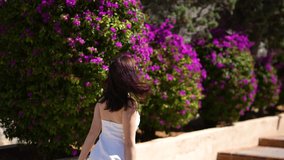 Girl in white dress is walking near the flower bushes in travel resort
