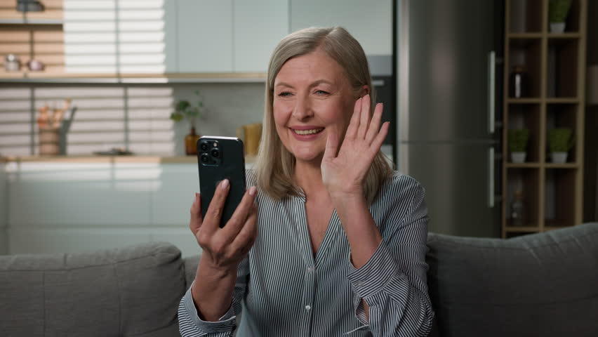 Happy elderly senior caucasian woman talking on video call using mobile phone waving hello smiling grandmother chatting at online conference by smartphone family chat at home sofa enjoy communication