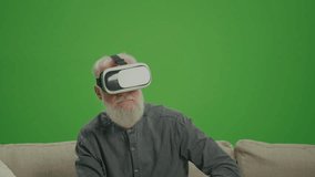 Green Screen. An Old Man With Gray Beard in VR Glasses Examines Everything Around, Sitting on a Sofa. Medical Applications of VR for the Elderly.