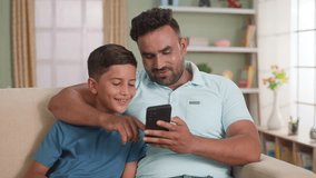 Happy smiling Indian father with son laughing by seeing mobile phone on sofa at home - concept of social media, weekend holidays and family time.