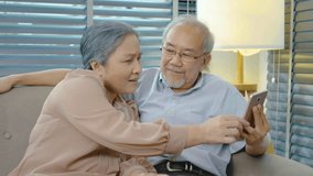 Happy elderly retired couple using a mobile phone together sitting sofa in the living room, using wireless internet connection, senior man and woman with smartphone, tease each other, slow motion