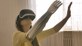 A woman in virtual reality glasses with a bionic arm prosthesis sits on the bed and moves her hands, looks around. The girl interacts with new technologies in a virtual digital universe.