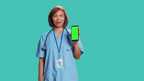 Clinic employee showing medical instructions video on phone green screen. Asian nurse wearing protective scrubs holding chroma key mock up phone, isolated over blue studio background