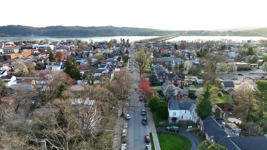 Drone view zooming in on a large community approaching a bridge over a beautiful river with hills in the background at sunset.