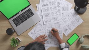 Close Up Top View Of Asian Male Artist Drawing Storyboard For The Film On The Table With Green Screen Smartphone And Laptop In The Studio
