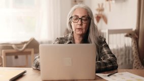 Portrait of beautiful gray haired senior woman with glasses having video call conference meeting by laptop computer at home Smiling mature female talking on working or personal conversation indoors