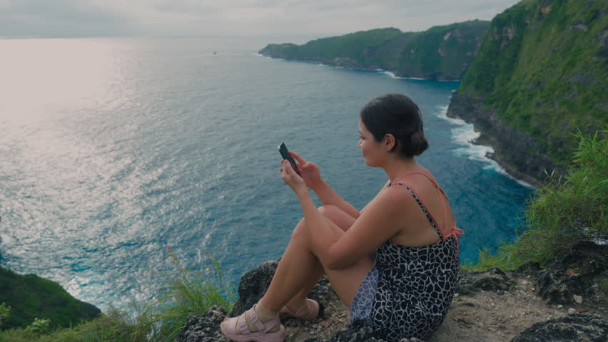 Asian girl relaxing on a rock above sea in summer with cell phone. Japanese woman holding hand phone sitting on stone at edge of cliff on ocean. Female hesitantly swipes his finger across phone screen