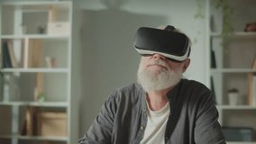 A Modern Old Man With Gray Beard in VR Glasses Examines Everything Around. An Elderly Man in VR Glasses Sitting at a Table at Home.VR to Help the Elderly.