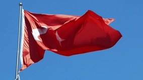 A large bright flag of Turkey flutters on a flagpole high above the ground against the backdrop of the sea, mountains and clouds.