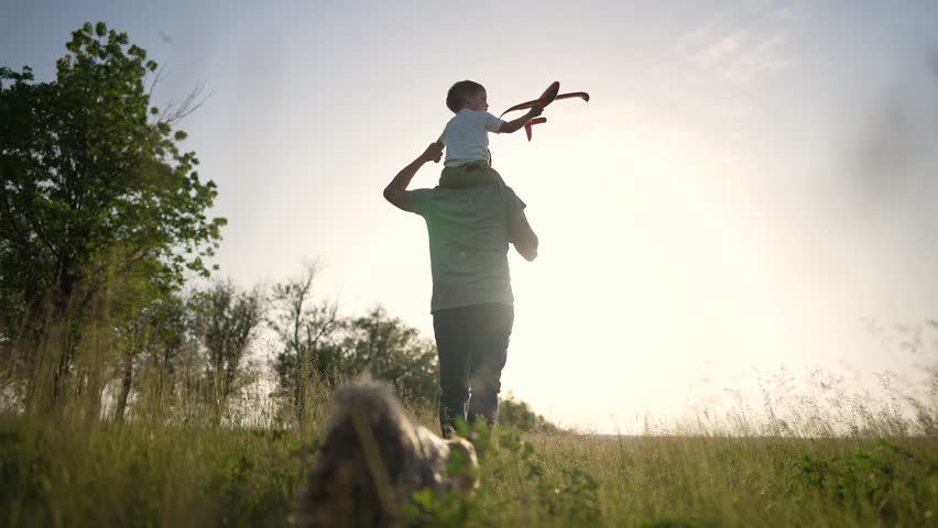 Happy family on walk. Son sits on father's neck. Boy is playing with a toy plane. Family walk with dog. The kid sits on the shoulders of his father and plays with plane. Walk in nature on green grass Royalty-Free Stock Footage #1105506211
