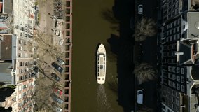 Aerial footage showcases the local architectural buildings and parked cars that line the river canal, while the tourist boat navigates the canal and passes under the bridge. Drone slowly ascends.