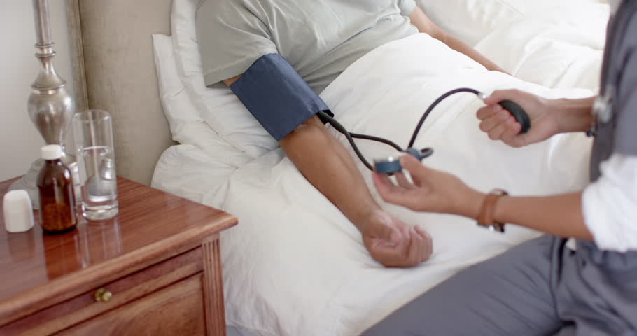 Diverse male doctor testing blood pressure of senior male patient in bed at home, slow motion. Medical services, check up, home visit, healthcare, wellbeing, and senior lifestyle, unaltered.