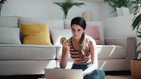 Video of confident woman using her laptop while eating an apple sitting on the floor at home.