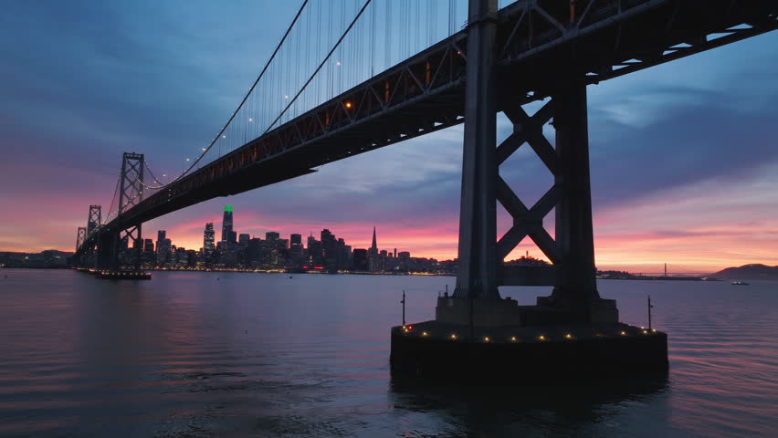 Drone flying low above calm water surface reflecting beautiful cloudy sky in pink and blue pastel colors. San Francisco downtown cityscape skyline seen under bridge in distance. Scenic night city view