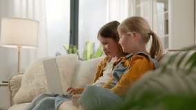 Two happy caucasian kids girls sisters friends children using laptop at home on couch looking at screen talking playing online games together on computer watching cartoon play on internet having fun