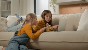 Two cute caucasian kids girls sisters friends play online games on laptop mobile phone playing together in living room on sofa older sister teaching little girl concept children and modern technology