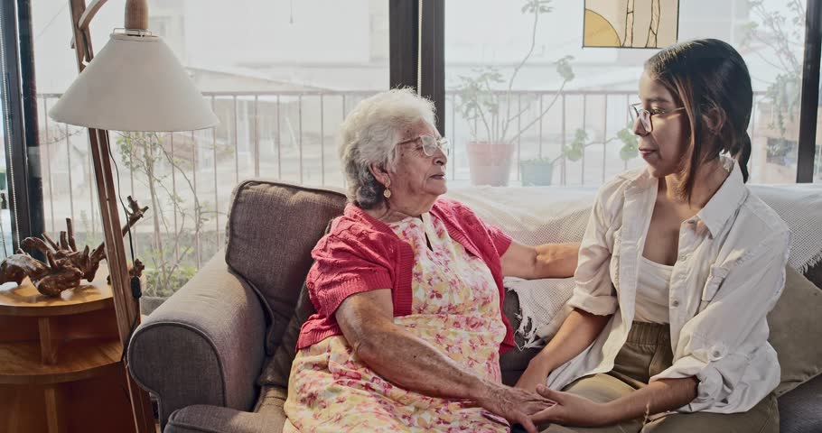 Grandmother and granddaughter sitting on the sofa at home, having a good talk, grandmother and granddaughter enjoying the moment together holding hands. Royalty-Free Stock Footage #1105517543