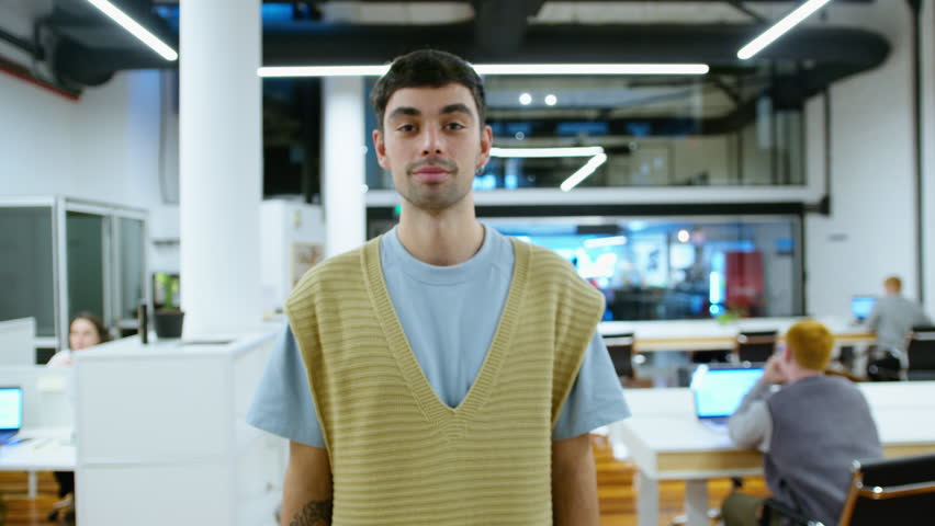 Young handsome businessman wearing earrings, knitted vest and casual t-shirt posing on camera with smile in modern open space office during the day. Video portrait, medium zoom shot