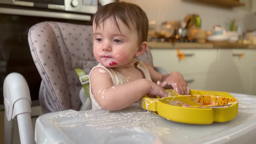baby girl eats dirty. happy family kid dream concept. baby funny got dirty in yogurt dairy product all over his face in drops learns to eat. baby eats dirty at home at the feeding lifestyle table Royalty-Free Stock Footage #1105523805