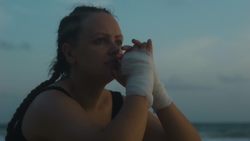 Sad female boxer MMA fighter on beach looks into distance with clasped hands. Royalty-Free Stock Footage #1105536723