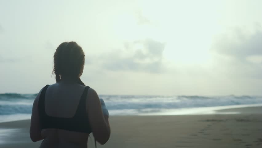 Female boxer MMA fighter girl boxing on beach at sunset sunrise. Back view.