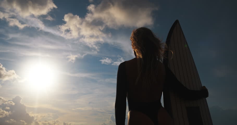 Dreamy romantic surfer woman enjoys amazing moment spent on sunny beach. Silhouette of woman dreamily looking into distance at setting sun, low angle rear view. Silhouette of woman with surfboard.