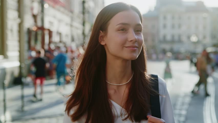 Shot of a confident young woman looking at the camera smiling and slowly walking poses on camera on a sunlight street. City life. Slow motion