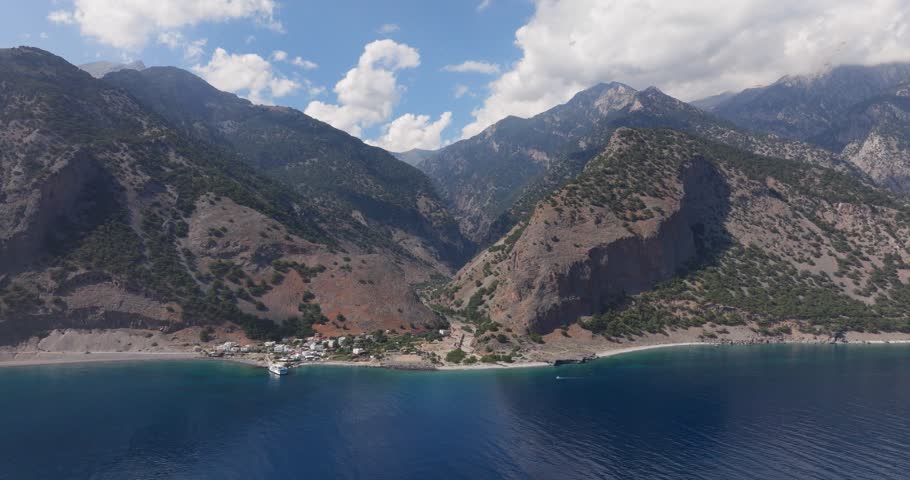 drone video of small fisherman town between mountains with blue turquoise clear sea and blue cloudy sky in crete agia roumeli