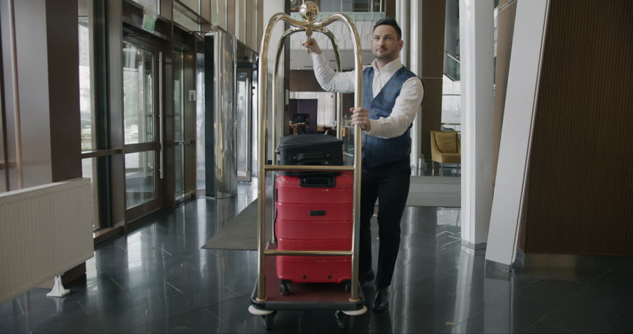 Hotel bellboy wearing uniform is walking along corridor pushing cart with luggage working alone. Porter occupation and guest service concept. Royalty-Free Stock Footage #1105547223