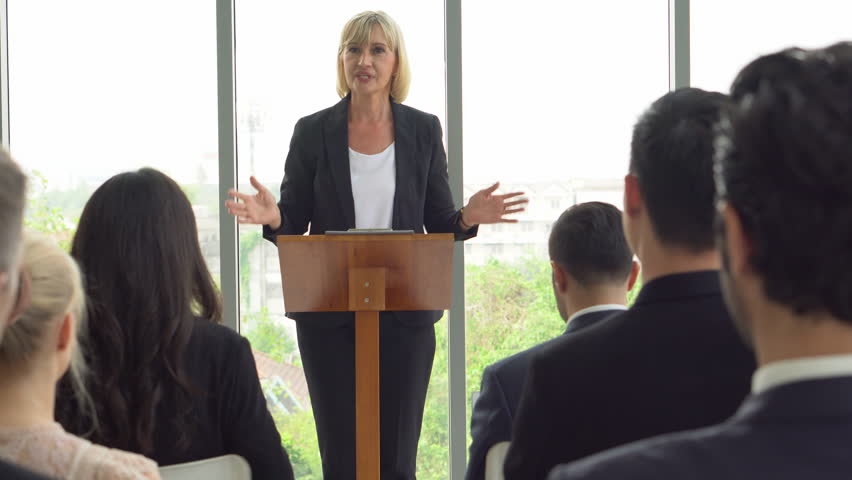 senior business woman standing at podium giving a speech addressing a conference in seminar. old executive female presentation in Meeting at office on window with colleagues laugh Royalty-Free Stock Footage #1105547581