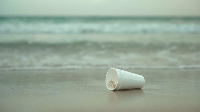 Close up of white foam cup on the sand on the seashore floating on the waves. People leave plastic waste and trash on the beach. Environment protection of nature