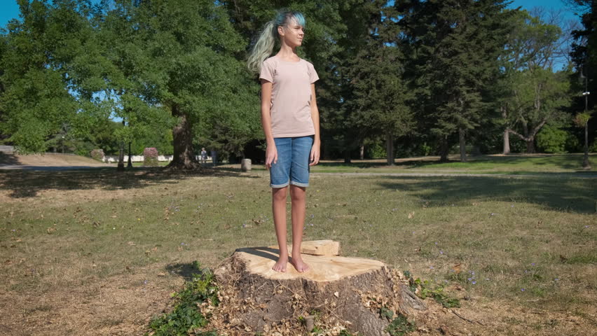 Smiling teen on stump. A view of happy modern teen girl stand on the stump in the green forest in summer. A concept of city weekend of children in the wood.