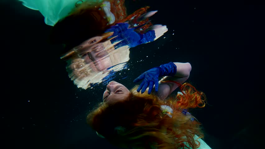 underwater magic, beautiful redhead woman floating under water surface and looking her reflection