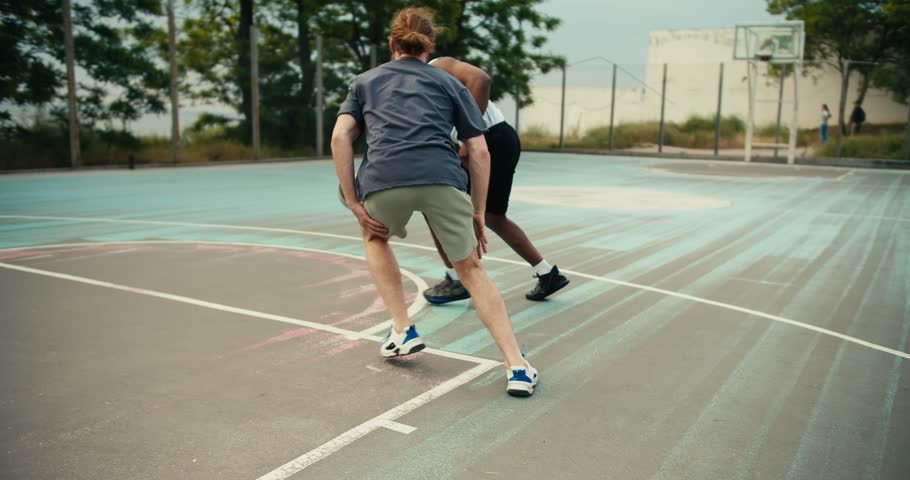 A Back man in a white t-shirt and a red-haired man in a gray t-shirt are playing basketball. The Black man outruns his opponent and scores a brilliant goal in a game of basketball Royalty-Free Stock Footage #1105573607