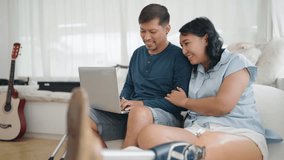 Happy asian couple using laptop for video call and waving hands to say hello. Woman in leg prosthesis and her husband having greeting through video call online with family on laptop