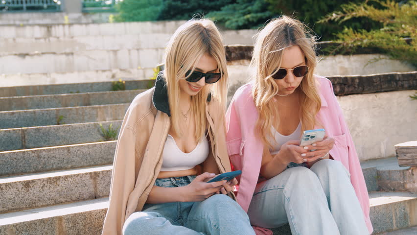 Two young beautiful smiling hipster female. Carefree women sitting at the stairs in the street in sunglasses. Models having fun. They looking at cellphone screen, using mobile apps, hold smartphone 