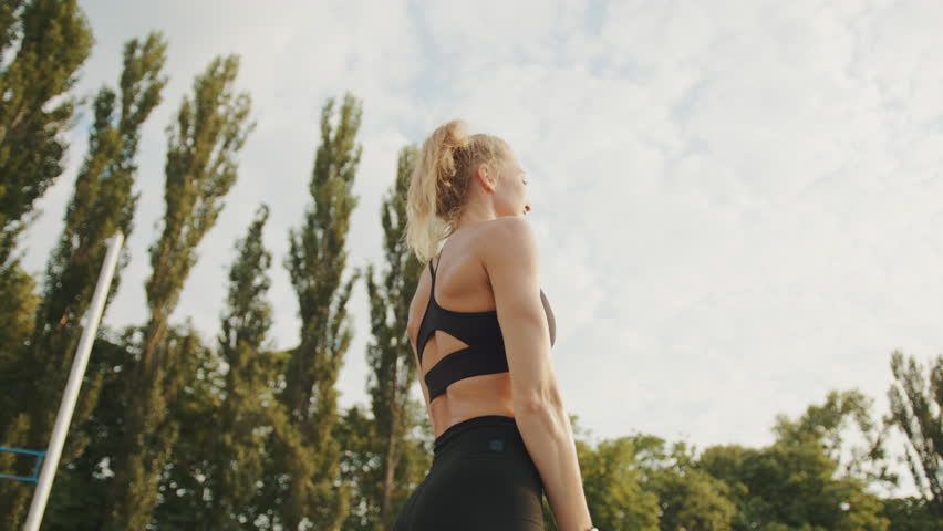 Medium shot of sporty blonde in black top performing arms stretching outside. Back view of young woman warming-up before running, sky and trees background. High quality 4k footage