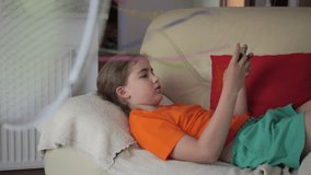 Girl Plays Video Games on Smartphone on Hot Heat Summer Day While Sitting on Sofa Near Fan. Child Plays Games on Mobile Phone in Heat Weather Near Fan. Cool Wind From Electric Fan at Summer Heat.