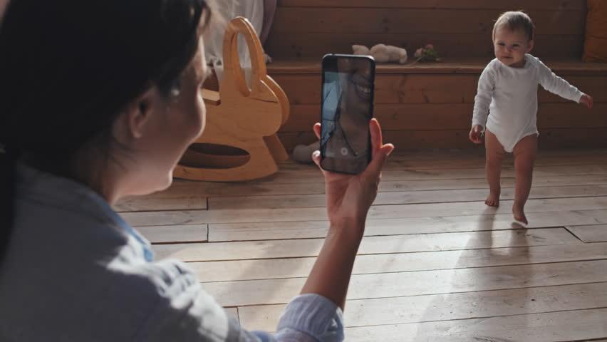 Over the shoulder shot of woman sitting on floor at home and making video of her barefoot baby making first steps with smartphone Royalty-Free Stock Footage #1105584053