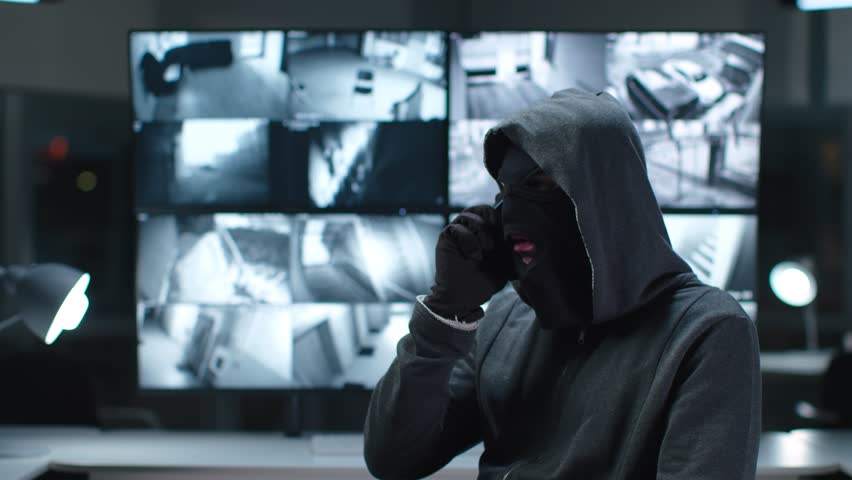 Criminal in mask sit in monitoring center with cctv footage and talking on phone. Hacker wearing balaclava talking on cellphone in video surveillance system room | Shutterstock HD Video #1105589105