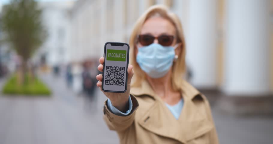 Mature woman in protective medical mask holding cellphone with electronic passport of vaccination looking at camera standing on city street | Shutterstock HD Video #1105589115