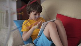 Boy Browsing Social Media on Tablet PC on Hot Heat Summer Day While Sitting on Sofa Near Fan. Child Plays Games on Tablet PC in Heat Weather Near Fan. Cool Wind From Electric Fan at Summer Heat.